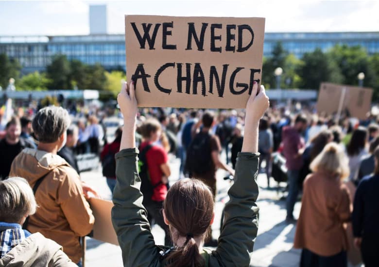 Photo of a protestor at a protest, holding a sign that reads 'We need a change'